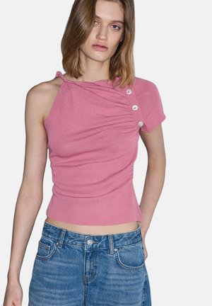 Mujer joven con un top asimétrico de manga corta rosa con botones y vaqueros azules de mezclilla sobre un fondo liso.