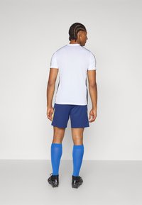 Hvid atletisk skjorte med sorte accenter, blå shorts og blå fodboldstrømper. Skjorten har korte ærmer, og outfittet inkluderer sorte fodboldstøvler.