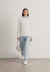 Biely pletený sveter cez svetlomodrú košeľu, skombinovaný so svetlomodrými džínsami a bielymi topánkami. V jednej ruke drží malú kabelku.