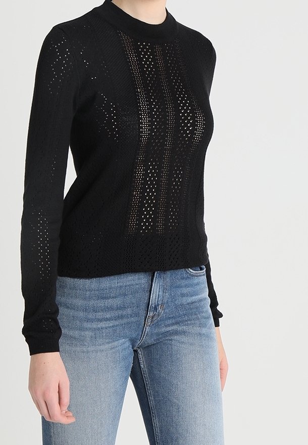 Schwarzer gestrickter Pullover mit langen Ärmeln, der offene Maschenmuster und einen hohen Kragen hat. Kombiniert mit hellblauen Jeans.