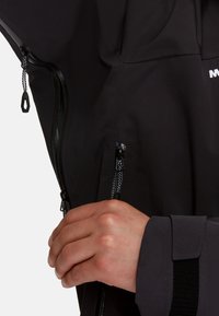 Giacca nera impermeabile con zip, caratterizzata da un tessuto testurizzato e polsini rinforzati. Primo piano di una mano che regola la zip.