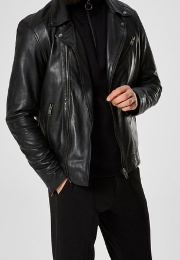 Man i en åtsittande svart läderjacka med dragkedjor över en svart topp, en hand i fickan, den andra handen höjd nära bröstet.