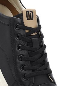 Sneaker in pelle nera con un tassello sul tallone beige con logo, design con lacci neri e occhielli rotondi. Superficie testurizzata visibile.