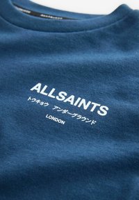 Tume-sinine dressipluus, mis on valmistatud pehmest puuvillast ning millel on ümmargune kaelus. Esiküljel on valge tekst "ALLSAINTS", mille kõrval on lisatekst jaapani keeles ning "LONDON."