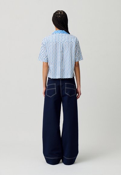 Camicia a maniche corte blu e bianca con motivo a pois, abbinata a pantaloni larghi blu scuro con cuciture bianche a contrasto e tasche posteriori.