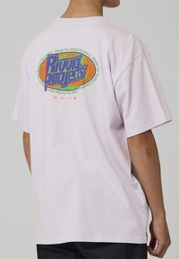 Lys lilla bomulls-T-skjorte med en flerfarget grafikk på baksiden med teksten "Riviera Projects" og flere design detaljer.