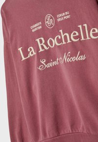 Bordo dukserica s vezanim tekstom "La Rochelle, Saint Nicolas" i dodatnom grafikom. Glatka tekstura, rebrasti pojas i rukavi.