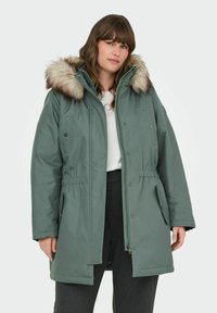 Zaļa parka jaka ar kažokādas oderi kapucē, elastīgu jostasvietu, spiedpogām un rāvējslēdzējiem, kā arī sānu kabatām. Gluda virsma ar matētu apdari.