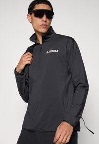 Melns četrtaugas rāvējslēdzēja krekls no viegla auduma ar elastīgām aprocēm, uz krūtīm baltā krāsā "TERREX" logotips un regulēšanas detaļas apakšmalā.