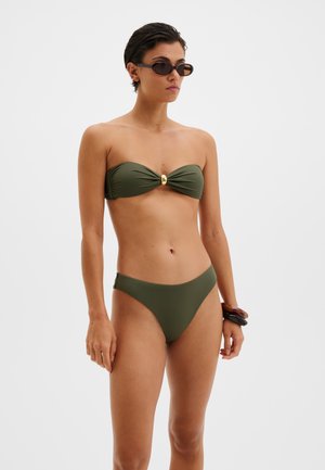 Femme portant un bikini bandeau vert olive avec un accent doré, des lunettes de soleil rectangulaires et des bracelets en écaille de tortue, debout devant un fond blanc uni.