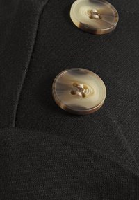 Černá látka se dvěma kulatými, mramorovaně hnědými knoflíky s texturovaným vzorem a čtyřmi dírkami pro upevnění.