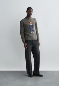 Pelēks adīts džemperis ar lapsas grafiku, apaļu dekoltē un ribotām aprocēm. Apvienots ar brīviem melniem džinsiem un melnajām kurpēm uz neitrāla fona.