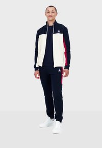 Survêtement bleu marine avec des accents crème, comprenant une veste zippée et un pantalon ajusté. Des baskets blanches complètent le look. Détail de logo brodé présent.