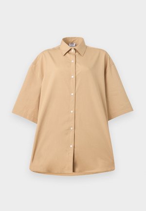 Egy nő bézs színű, túlméretezett rövid ujjú ingben és hozzáillő rövidnadrágban, fekete kézitáskát tart, barna szandált visel, fehér háttér előtt áll.