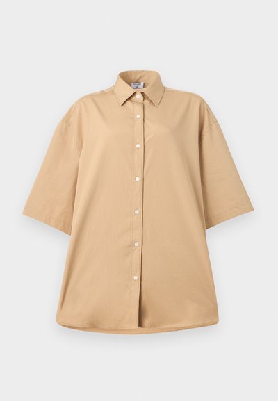 Μπεζ oversized πουκάμισο με κοντά μανίκια, μυτερό γιακά και λευκά κουμπιά μπροστά, σε απλό λευκό φόντο.