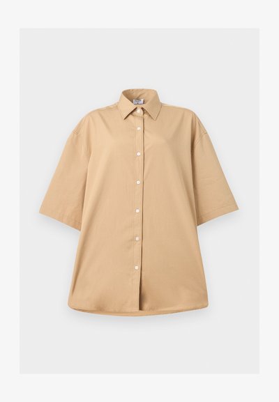 Μπεζ oversized πουκάμισο με κοντά μανίκια, μυτερό γιακά και λευκά κουμπιά μπροστά, σε απλό λευκό φόντο.