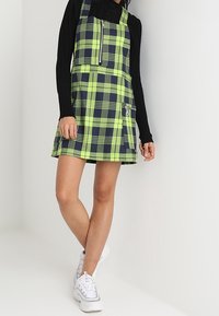 Karierte Latzkleid in Limettengrün und Marineblau, mit einem Reißverschluss vorne, seitlicher Tasche und schwingendem Saum; kombiniert mit einem schwarzen Langarmoberteil und weißen Sneakers.