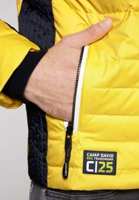 Żółta kurtka ocieplana z zamkiem błyskawicznym, posiadająca kieszeń z czarnym zamkiem i naszywką z logo, kontrastujący czarny materiał po bokach, teksturowana powierzchnia.