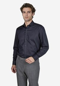 Donkerblauwe overhemdd met lange mouwen, gladde textuur en getailleerd ontwerp. Gedragen met grijs geruite broek.