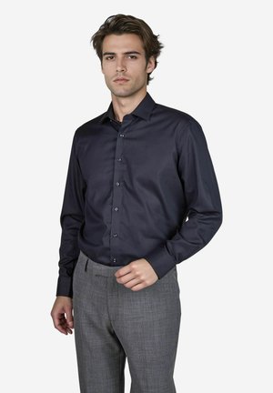 Dunkelblaues Hemd mit langen Ärmeln, glatter Textur und figurbetontem Design. Trägt eine graue Karohose.