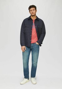 Marineblaue Jacke mit Knöpfen und zwei Brusttaschen über einem roten T-Shirt, kombiniert mit hellblauen Jeans und weißen Sneakers. Schlichte Texturen, lässige Passform.