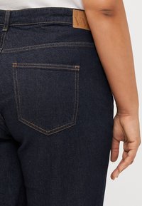 Dark denim jeans are shown from the back, highlighting a single pocket and a tan brand label. A hand rests casually on the hip.