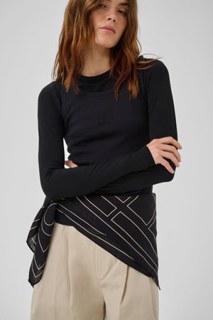 Femme aux cheveux châtains mi-longs portant un haut noir à manches longues et un pantalon beige, avec une écharpe noire à motifs nouée autour de sa taille.