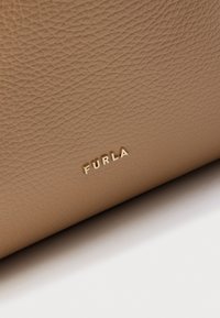 Geantă bej din piele texturată, cu logo-ul auriu „FURLA” în partea de jos. Suprafața are un finisaj granulă și este ușor lucioasă.