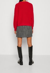 Maglione rosso lavorato a maglia con maniche a palloncino, abbinato a una gonna a scacchi bianco e nero e stivali neri fino al ginocchio. Dettagli del tessuto texture visibili.