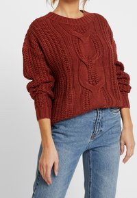 Gruby sweter z dzianiny w kolorze rdzy z fakturowanymi wzorami warkoczy. Posiada okrągły dekolt i oversize'owe rękawy. Połączony z niebieskimi jeansami.