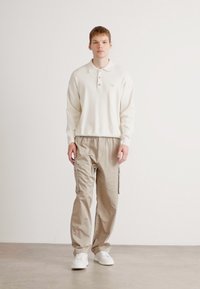 Tricou polo alb cu trei butoane, asortat cu pantaloni cargo bej cu buzunare laterale și încălțăminte albă. Textură netedă, croială lejeră.