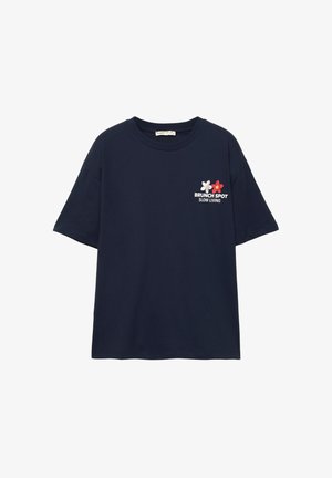 Laivansinis puuvillapaita, jossa on lyhyet hihat ja pyöreä kaula-aukko. Edessä on värikäs kukkakuva ja teksti: "BRUNCH SPOT SLOW LIVING."