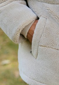 Jachetă din fleece bej cu o suprafață texturată, având un buzunar lateral, manșete ribbed și o croială lejeră. Material moale, călduros, vizibil.