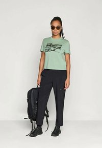 Zaļš kokvilnas T-krekls ar grafisko apdruku, apvienots ar melniem biksēm ar rāvējslēdzējiem. Modelis tur melnu mugursomu un valkā saulesbrilles.