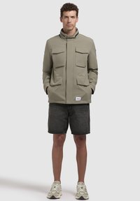 Jachetă khaki cu guler înalt, două buzunare mari pe piept și buzunare laterale. Asortată cu pantaloni scurți negri și adidași de culoare deschisă.