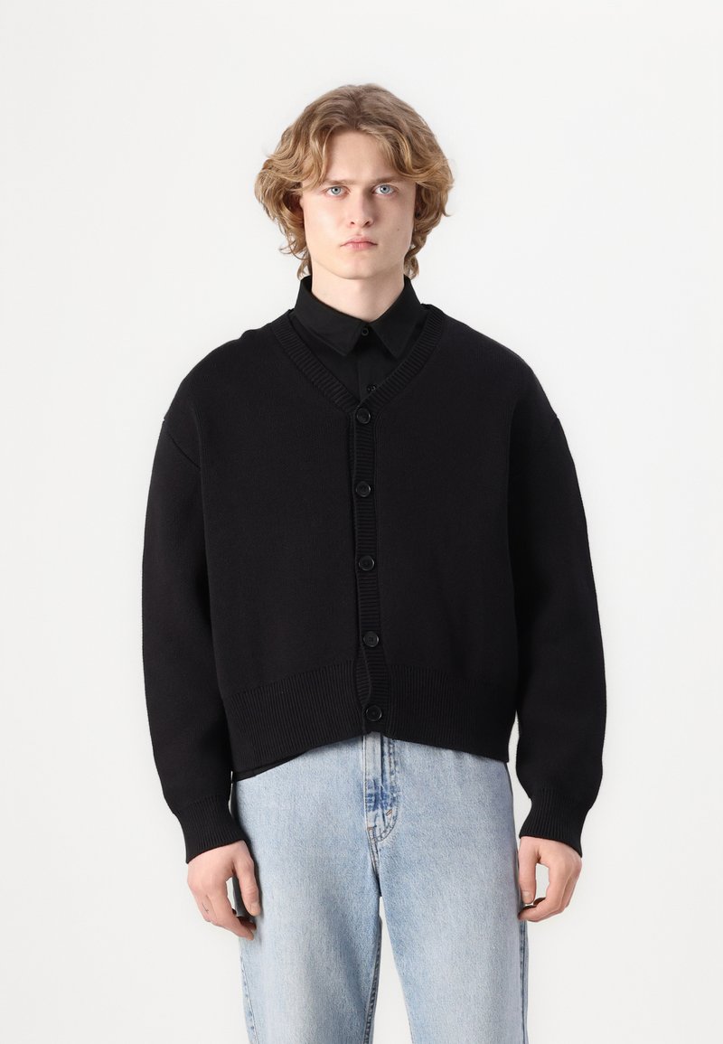 Cardigã preto de malha com decote em V, botões à frente e bordas caneladas. Usado sobre uma camisa preta e combinado com jeans de tom azul claro.