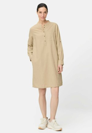 Kvinde stående iført en beige langærmet skjortekjole og hvide sneakers, hænderne afslappede, neutral baggrund.