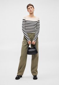 Top de mangas longas com riscas em creme e azul marinho, combinado com calças largas verde-oliva. Modelo segura uma bolsa preta de palha; sapatos pretos robustos.