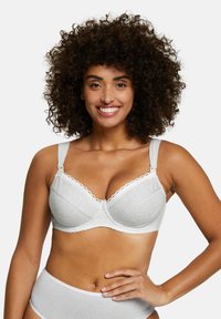 Femme souriante aux cheveux bouclés portant un soutien-gorge gris clair orné de dentelle et une culotte assortie, posant avec une main sur la hanche.