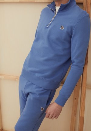 Hombre con sudadera azul con cremallera parcial y pantalones cortos a juego con pequeño logo de cebra, apoyado contra un marco de madera sobre fondo beige.