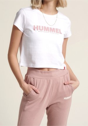 Nainen valkoisessa lyhytvartisessa T-paidassa ja vaaleanpunaisissa collegehousuissa, joissa on "HUMMEL"-logo, kädet taskuissa, beige tausta.