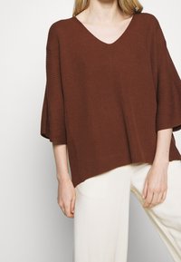 Top marrone a coste con scollo a V e maniche ampie, abbinato a pantaloni leggeri di colore crema, morbidi e dal taglio largo.