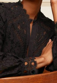 Blusa nera in pizzo con intricati motivi floreali, scollatura alta, apertura a chiave e rifiniture decorative sulle maniche con ritagli circolari.