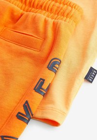 Orange bomuldsjoggingbukser med en struktureret talje, en baglomme og en mørkeblå trykt logoaccent langs kanten.