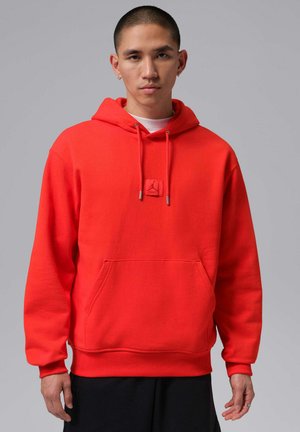 Junger Mann, der einen knallroten Hoodie mit Fronttasche und schwarze Hose vor einem schlichten grauen Hintergrund trägt.