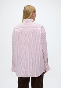 Γυναίκα με μακριά ξανθά μαλλιά που φοράει άνετο λευκό πουκάμισο με κόκκινες κάθετες ρίγες, εμφανιζόμενη από την πίσω όψη σε απλό φόντο.