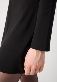 Vestido preto de malha com mangas compridas e uma fenda lateral. O tecido tem um acabamento texturizado, e a bainha apresenta um detalhe de costura simples.