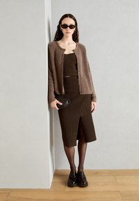 Robe marron à côtes avec une fente jusqu'aux genoux, superposée sous un cardigan marron duveteux. Le mannequin tient un petit sac noir ; des lunettes de soleil foncées sont portées.