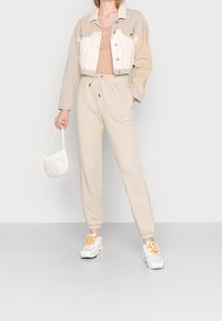 Korthällt tvåfärgat denimjacka med silvriga knappar, beige joggers med dragsko och Nike-logga, samt vita sneakers med orange detaljer.