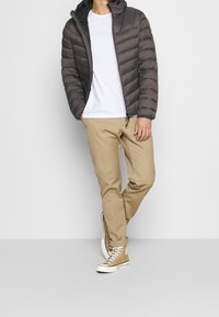 Polstret mørkegrå jakke med hætte, hvid t-shirt og beige bukser. Brune høje sneakers med hvide såler og snørebånd.
